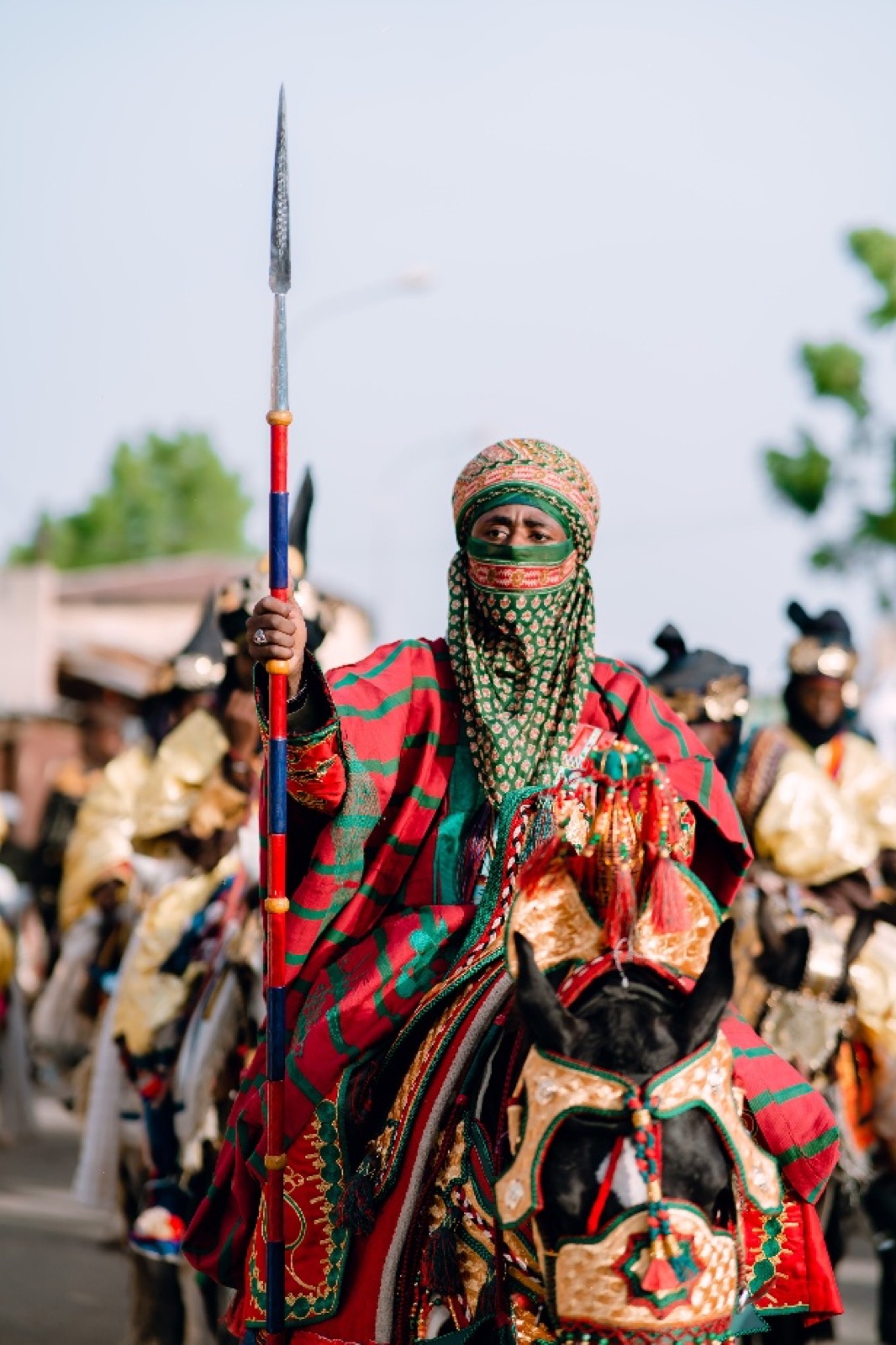 A rider in colourful ceremonial clothing holds a long spear while riding a decorated horse, with other riders behind him.